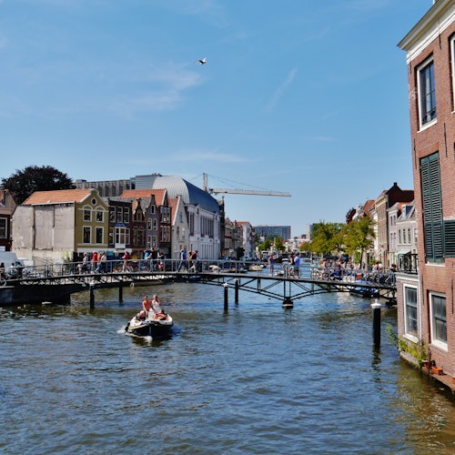 A canal with a boat, a bridge with pedestrians, historic buildings, and a crane in the background under a clear sky.