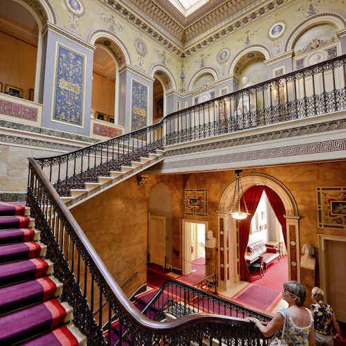 Dvije osobe pored veličanstvenog stubišta s raskošnim ogradama, crvenim tepihom, visokim lukovima i ukrasnim zidovima u povijesnom zdanju.