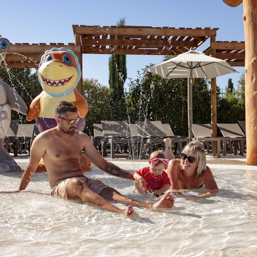 A man, woman, and child play in a shallow pool with water jets, surrounded by cartoon character statues and lounge chairs.