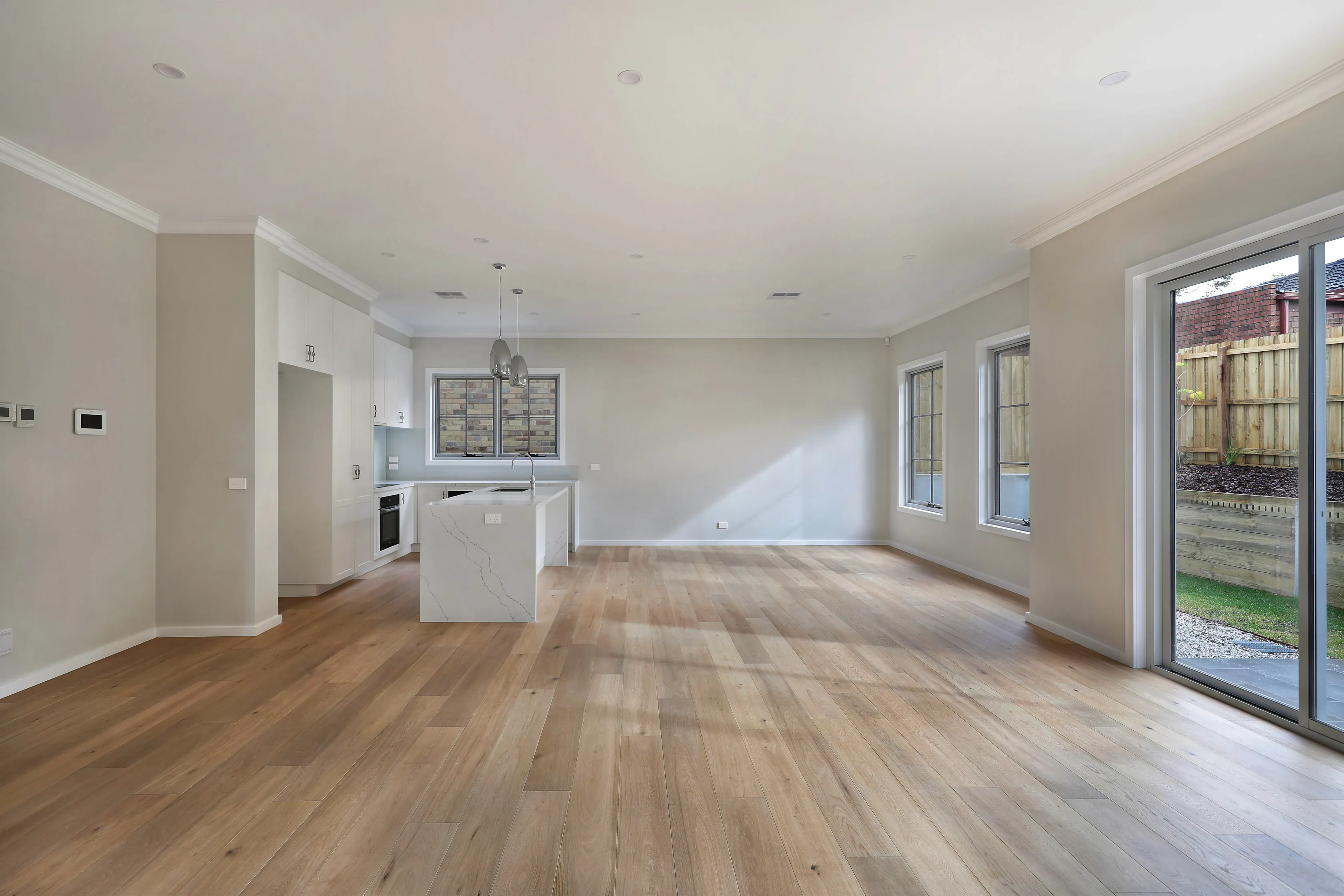 Empty open-plan living and dining area with warm oak floors, a marble waterfall island in the white kitchen and large sliding doors opening to a fenced backyard