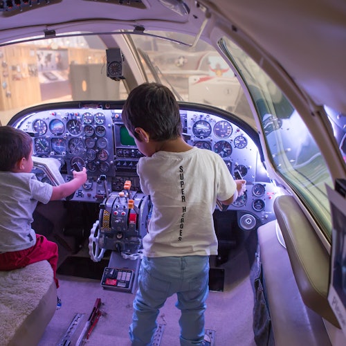 Two young children in a cockpit, one sitting and one standing, both interacting with the aircraft's controls.