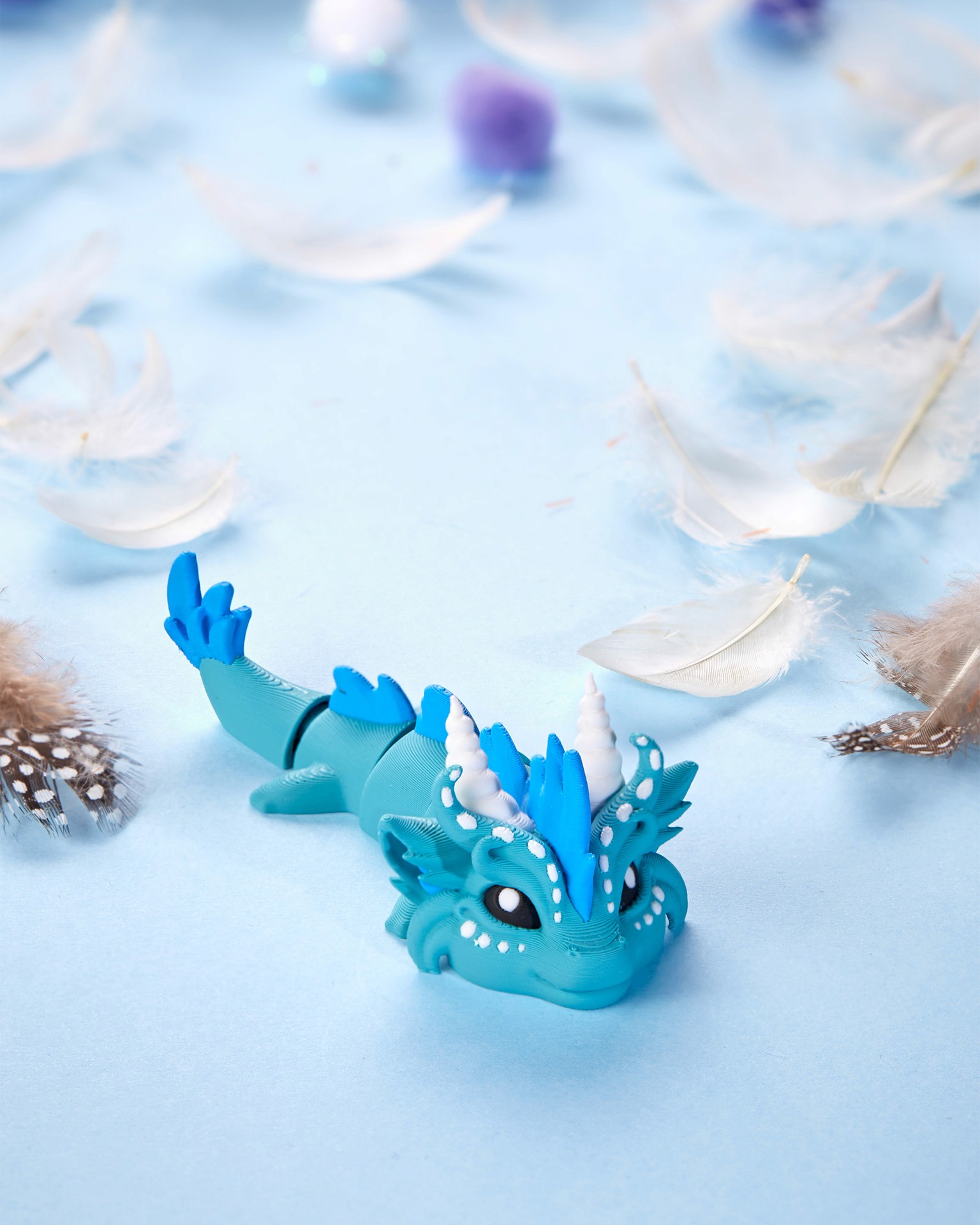 A small, intricately detailed blue dragon figurine lies on a light blue surface, surrounded by scattered white and brown feathers, showcasing creative 3D print artistry.