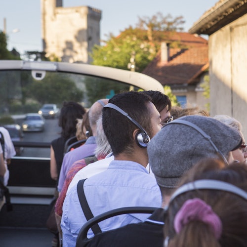 Lidé se sluchátky na uších sedí na otevřeném autobuse prohlídkového okruhu, který projíždí městem s budovami a vzdálenou věží.