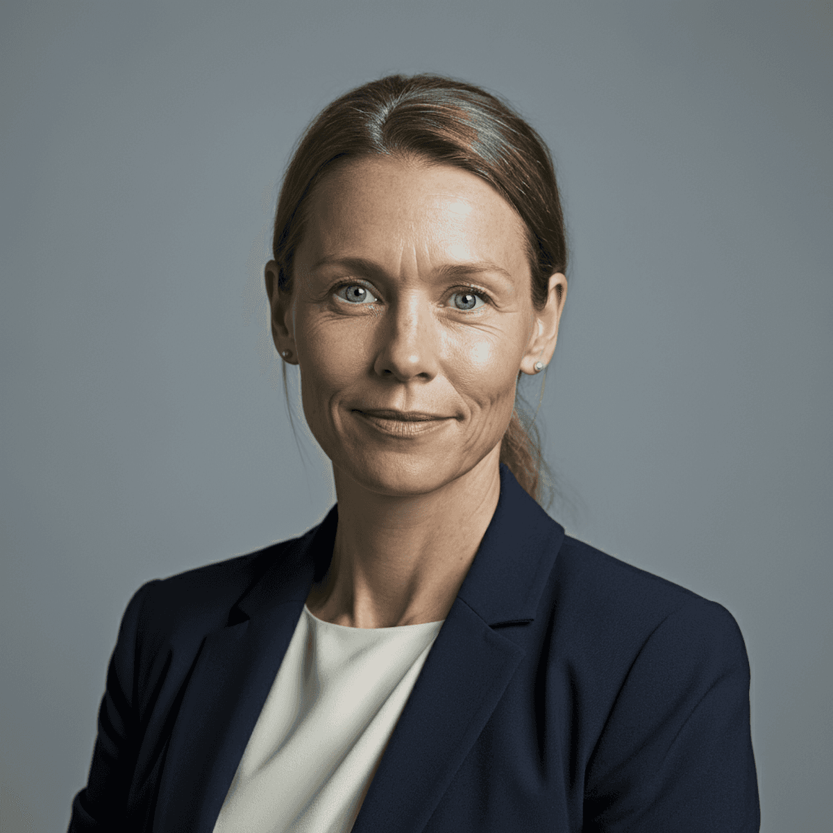 Professional woman with light brown hair in a ponytail, wearing a navy blazer and white blouse. She has a confident and calm expression, set against a gray background.