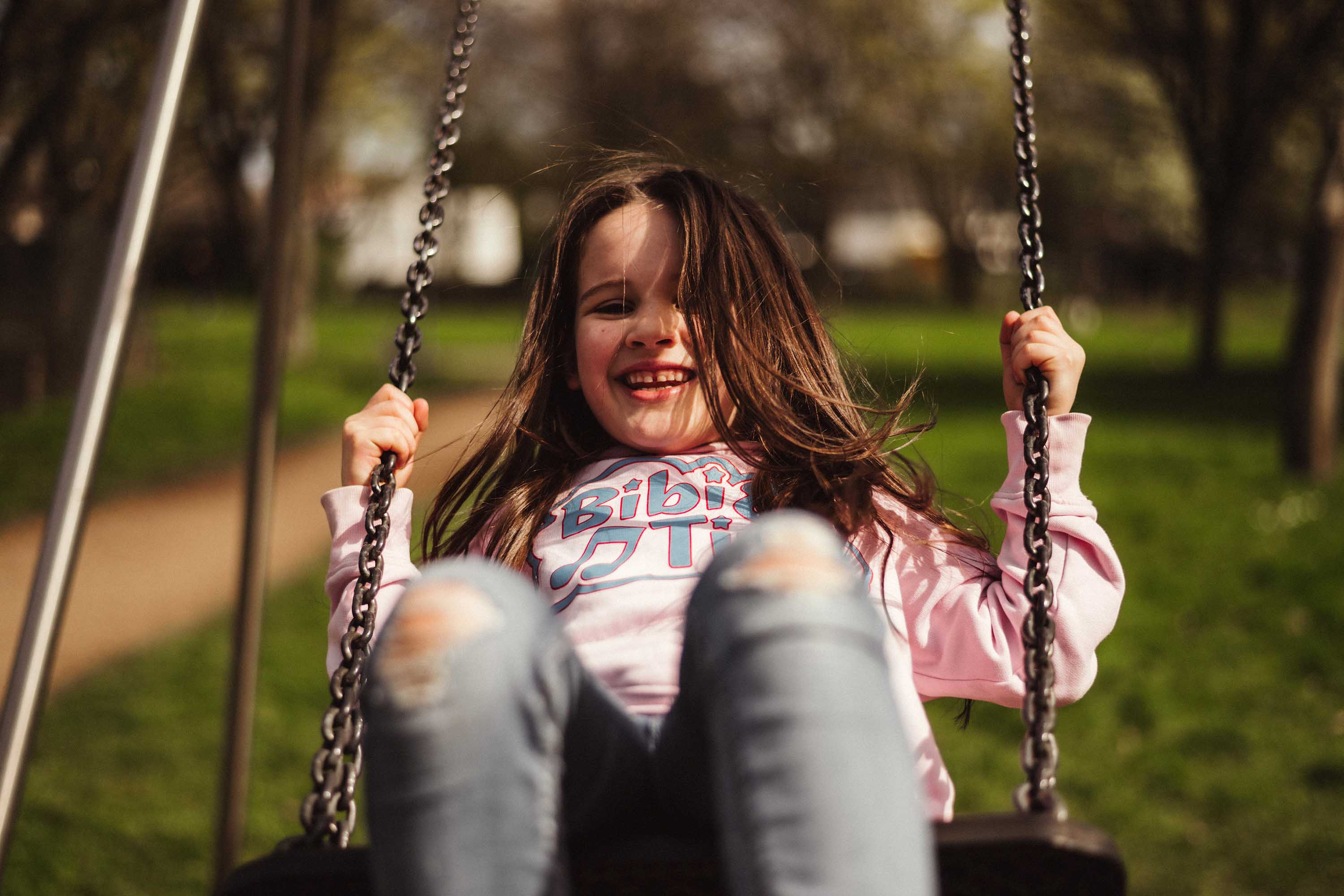 Authentische Kindergartenfotografie Gevelsberg: Ein Mädchen auf einer Schaukel schwingt hoch in die Luft, fotografiert von PK photography 4 Kids NRW. Der Hintergrund zeigt unscharfe Bäume und ein weiches Bokeh