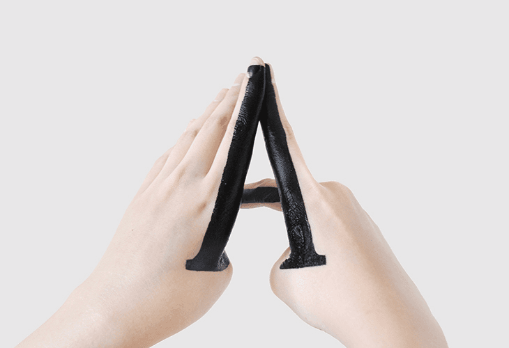 Two hands are shown holding black fabric strips against a light gray background. The strips are folded in half and held between the fingers. The fabric appears soft and flexible. The hands are positioned centrally in the frame, emphasizing the material's texture and form.