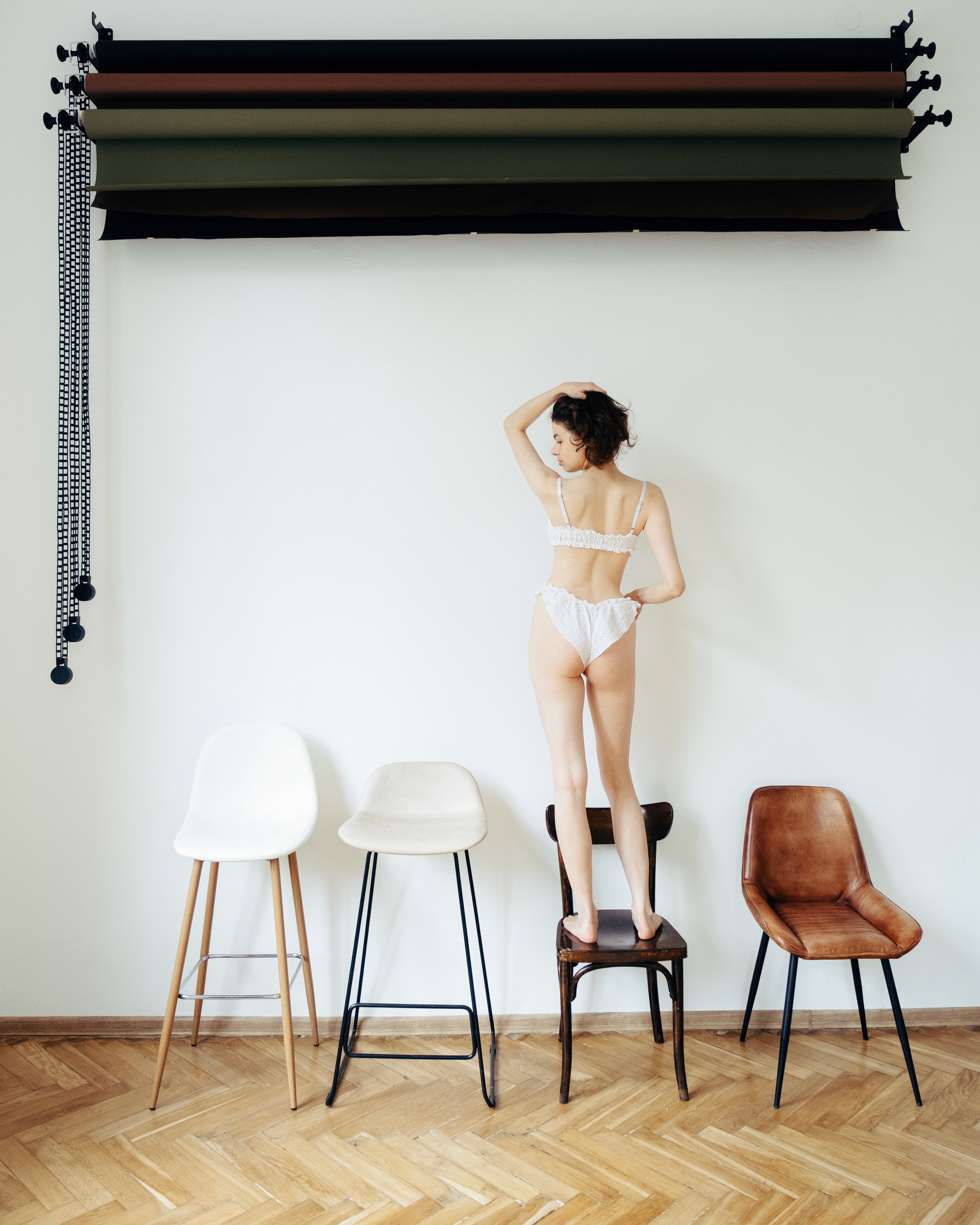 Palina – Soft Laziness – standing on a chair beneath hanging backdrops, looking away in delicate lingerie