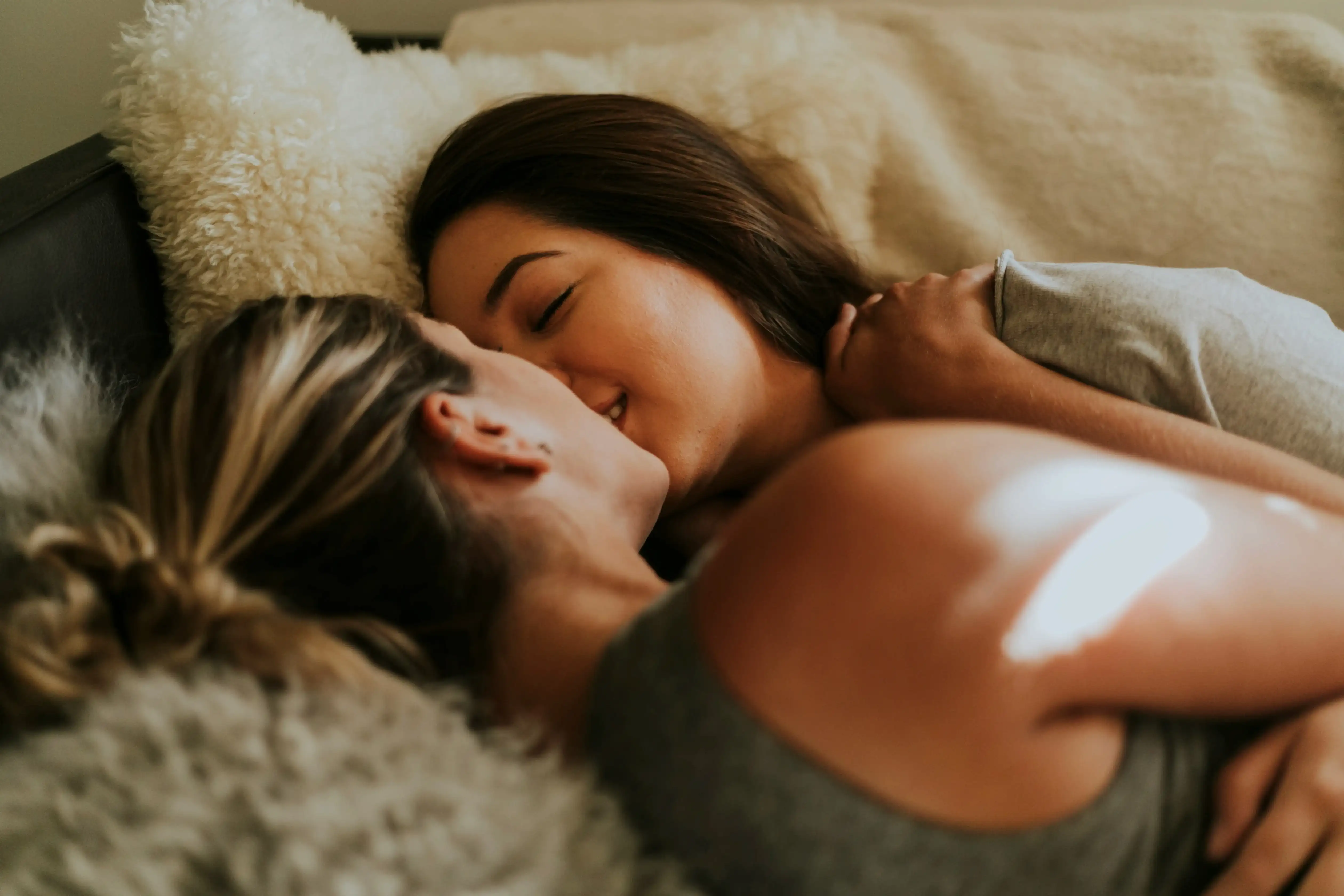 Two women lie close together on a soft bed, gently kissing and embracing, conveying intimacy, trust, and emotional connection—reflecting themes of queer pleasure, safety, and being present in one’s body.