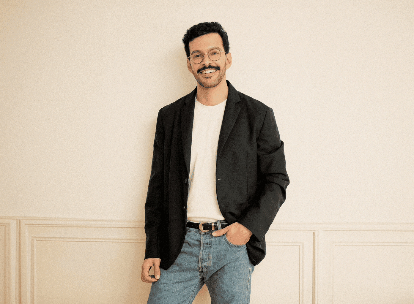 A portrait of a man standing casually in a blazer, jeans, and glasses — confident, relaxed, and approachable with a natural warmth.