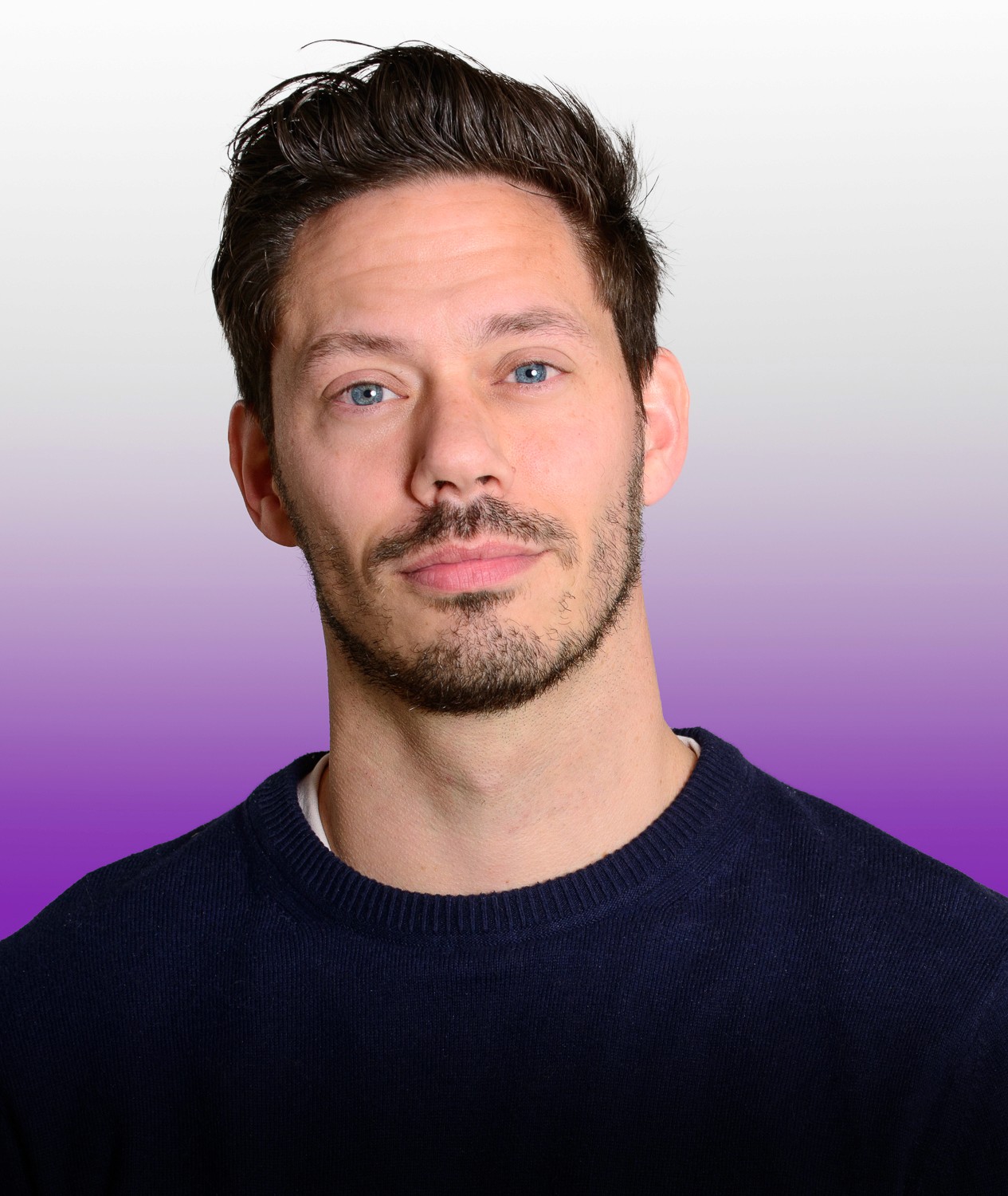 A young person with curly hair smiles at the camera while sitting in front of a laptop against a dark background.