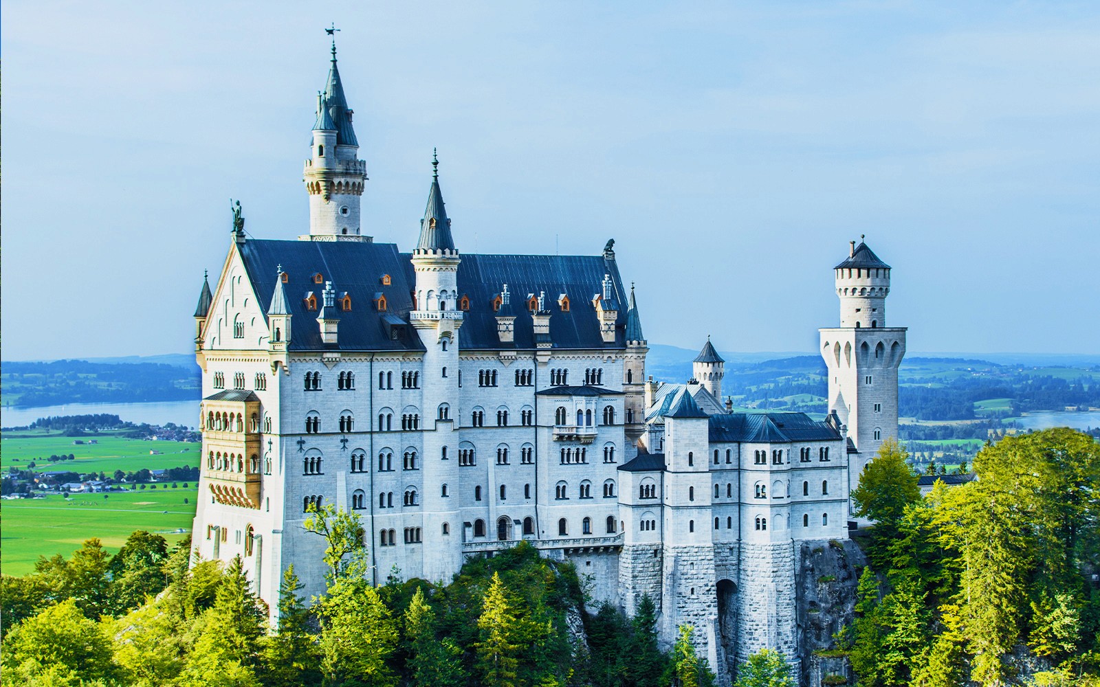 Neuschwanstein Castle in Bavaria, Germany, surrounded by lush greenery and distant hills.