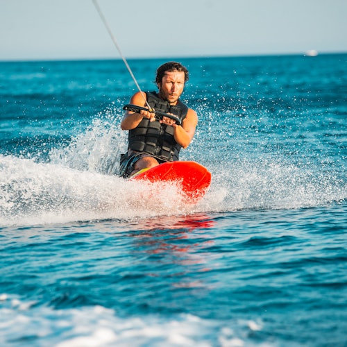 A person in a black life vest is wakeboarding on blue water, holding a handle attached to a rope.