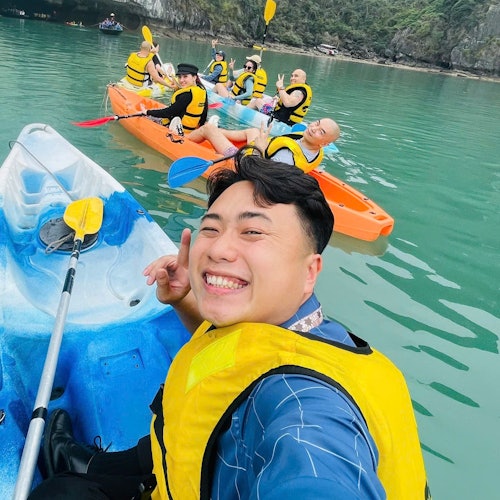 En smilende person i en gul redningsvest tager en selfie på en blå kajak, mens en gruppe mennesker kajakker i baggrunden på en rolig sø.