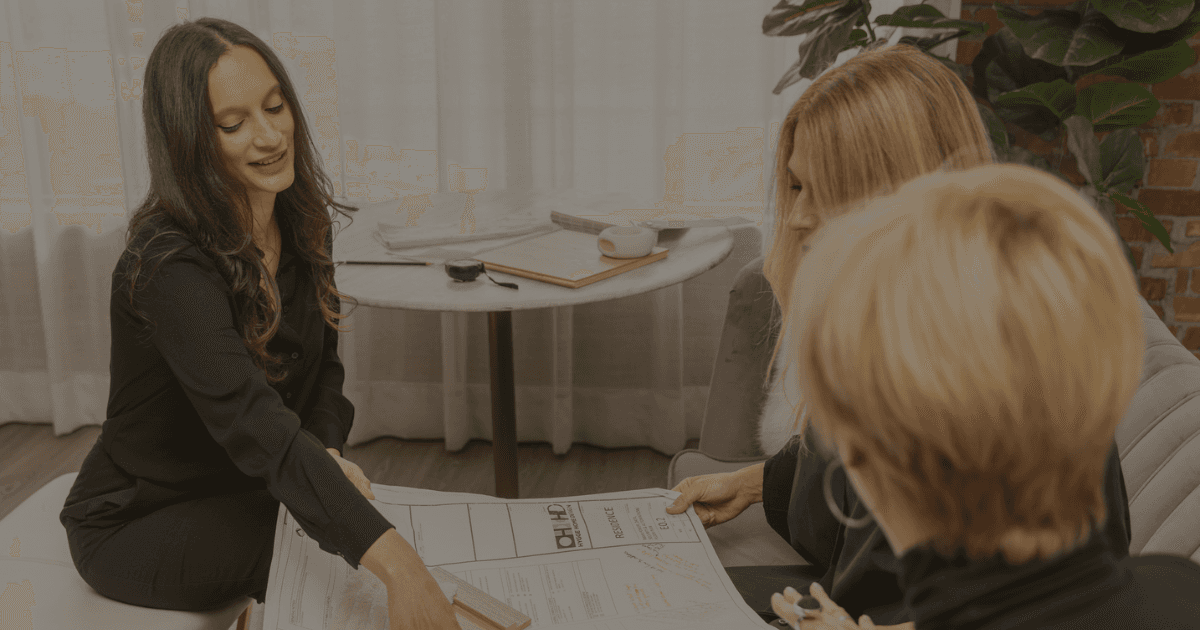 A dark haired woman points to a drawing that two blond fair skinned women are holding. It appears she is trying to explain a part of the design.