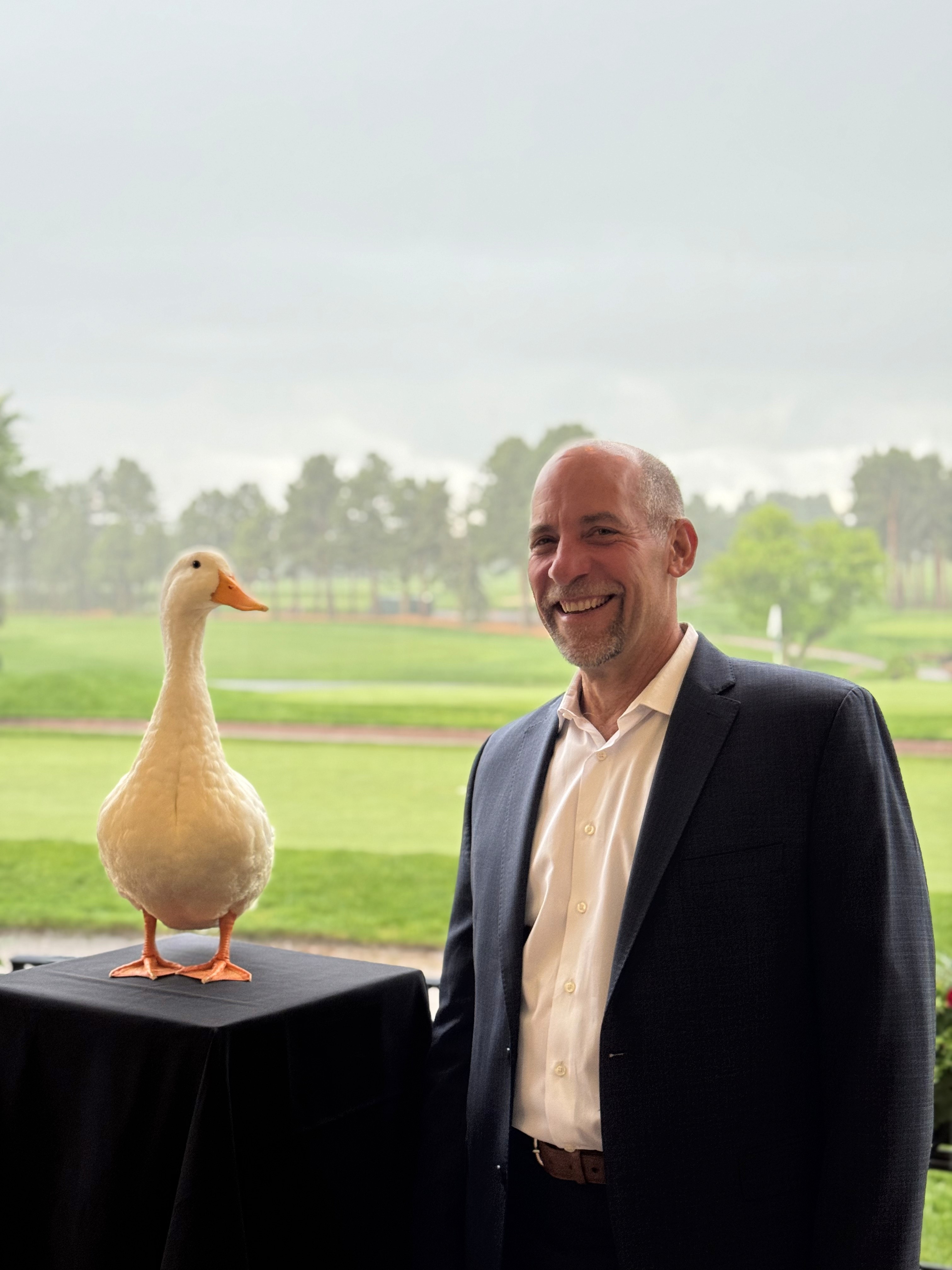 John Smultz in a suit stands smiling next to the Aflac duck perched on a black-covered table, with a green golf course and trees visible in the background.