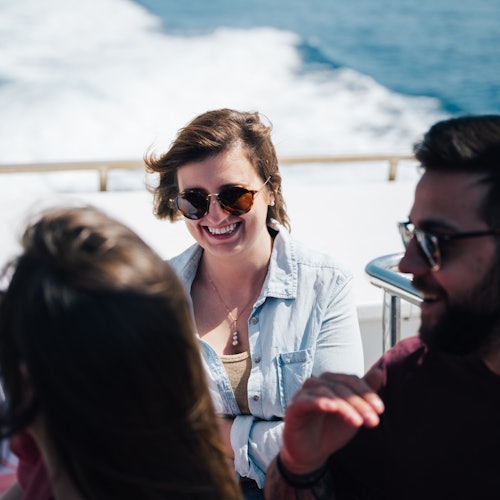 Tři lidé si užívají jízdu na lodi, přičemž dva z nich se usmívají a mají sluneční brýle. V pozadí je vidět oceán.