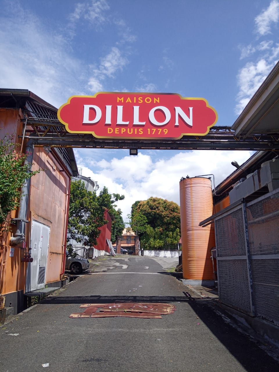 Entrée de la distillerie Maison Dillon, fondée en 1779, avec une enseigne rouge vif. Flanquée de bâtiments industriels et d'une végétation luxuriante sous un ciel bleu.