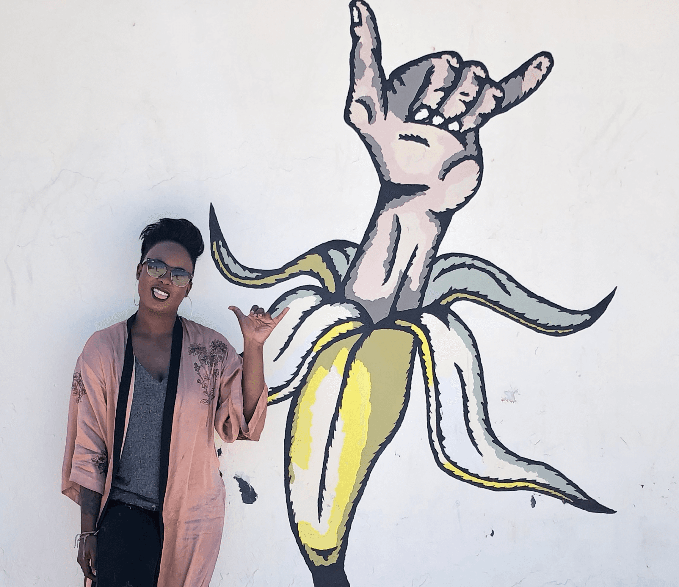 A person stands next to a colorful mural of a banana with a hand making a gesture on a white wall.