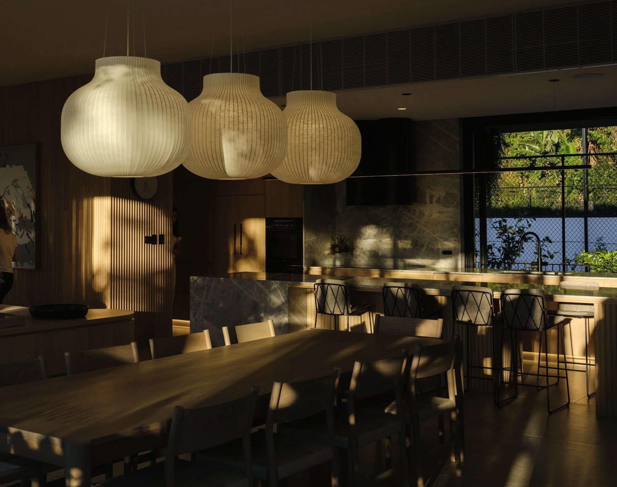 Warm timber kitchen and dining space with marble island, ribbed cabinetry, and sculptural pendant lights in evening light.