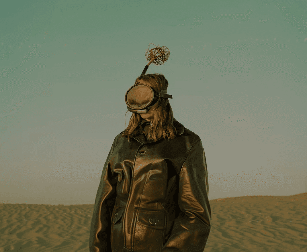 A person wearing a mask with an antenna standing against a pale sky.