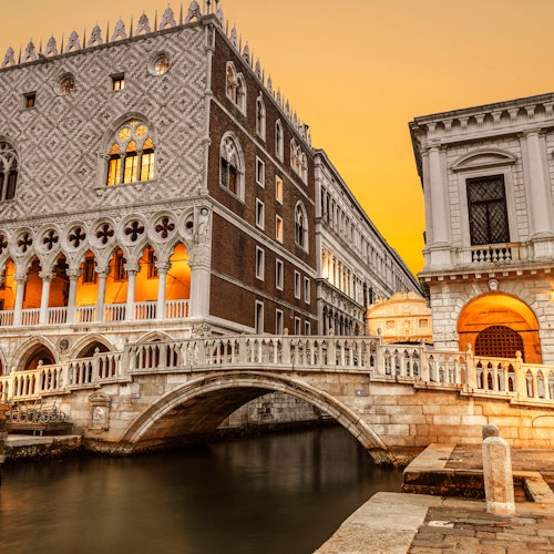 Edifici storici con facciate ornate e archi sono collegati da un ponte sopra un canale al tramonto.