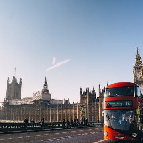 Red double-decker bus on a bridge, with the Houses of Parliament and Big Ben in the background under a clear sky.