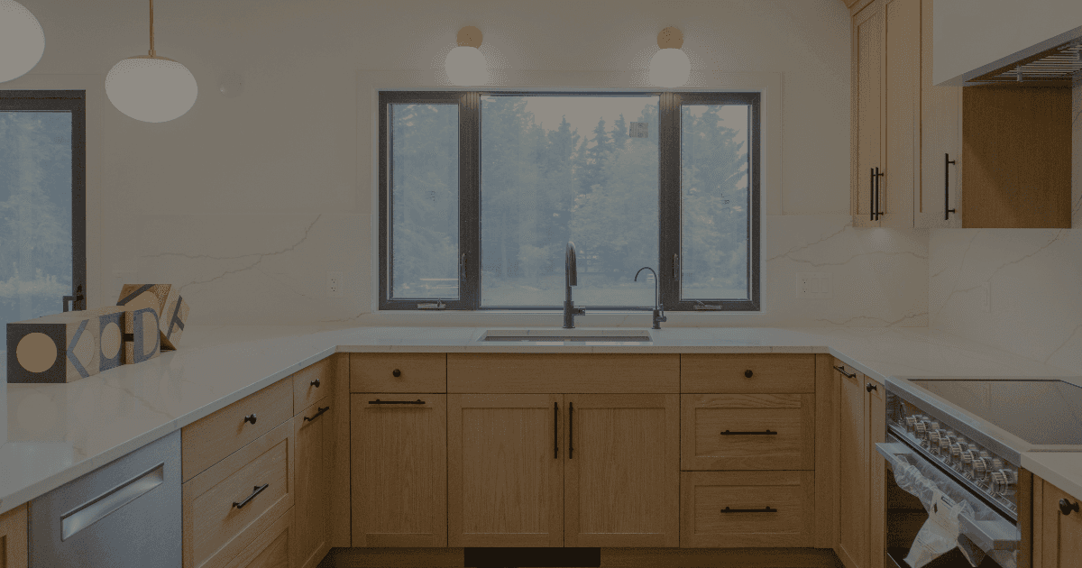 A neutral light wood kitchen in a U shape design. Facing a kitchen sink in front of a large window with black casings.