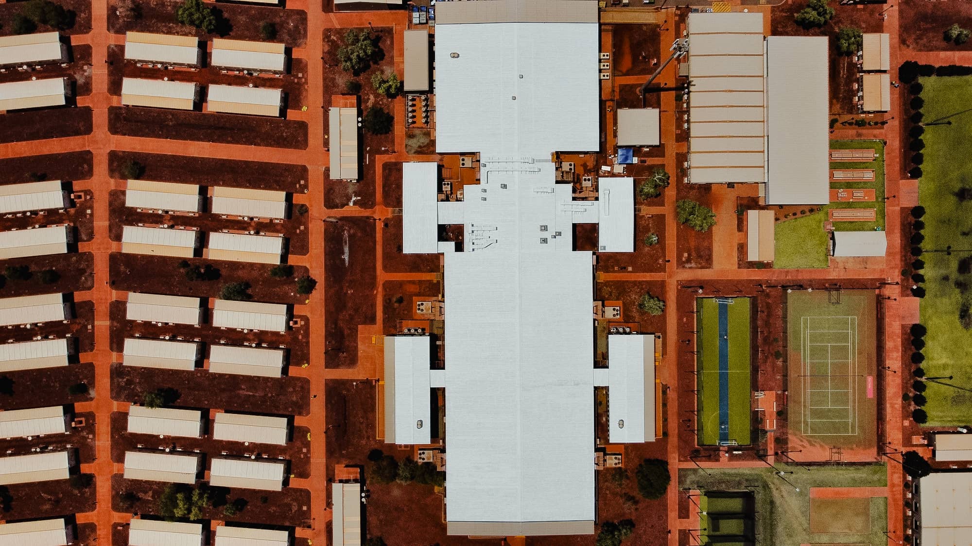 Aerial view of a mining camp with accommodation blocks, large industrial buildings, sports courts, and red earth terrain in Pilbara, Western Australia