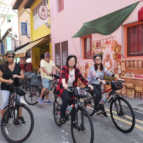 สี่คนกำลังขี่จักรยานบนถนนที่เต็มไปด้วยร้านค้า หนึ่งในกำแพงมีภาพวาดหลากสีสันแสดงอาหารต่าง ๆ และมีผู้ขายอยู่ด้วย