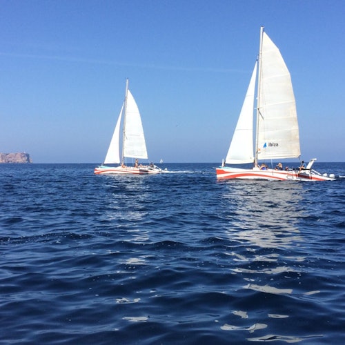 Två segelbåtar med vita segel rör sig över ett lugnt blått hav under en klar himmel, med avlägsen land synlig vid horisonten.