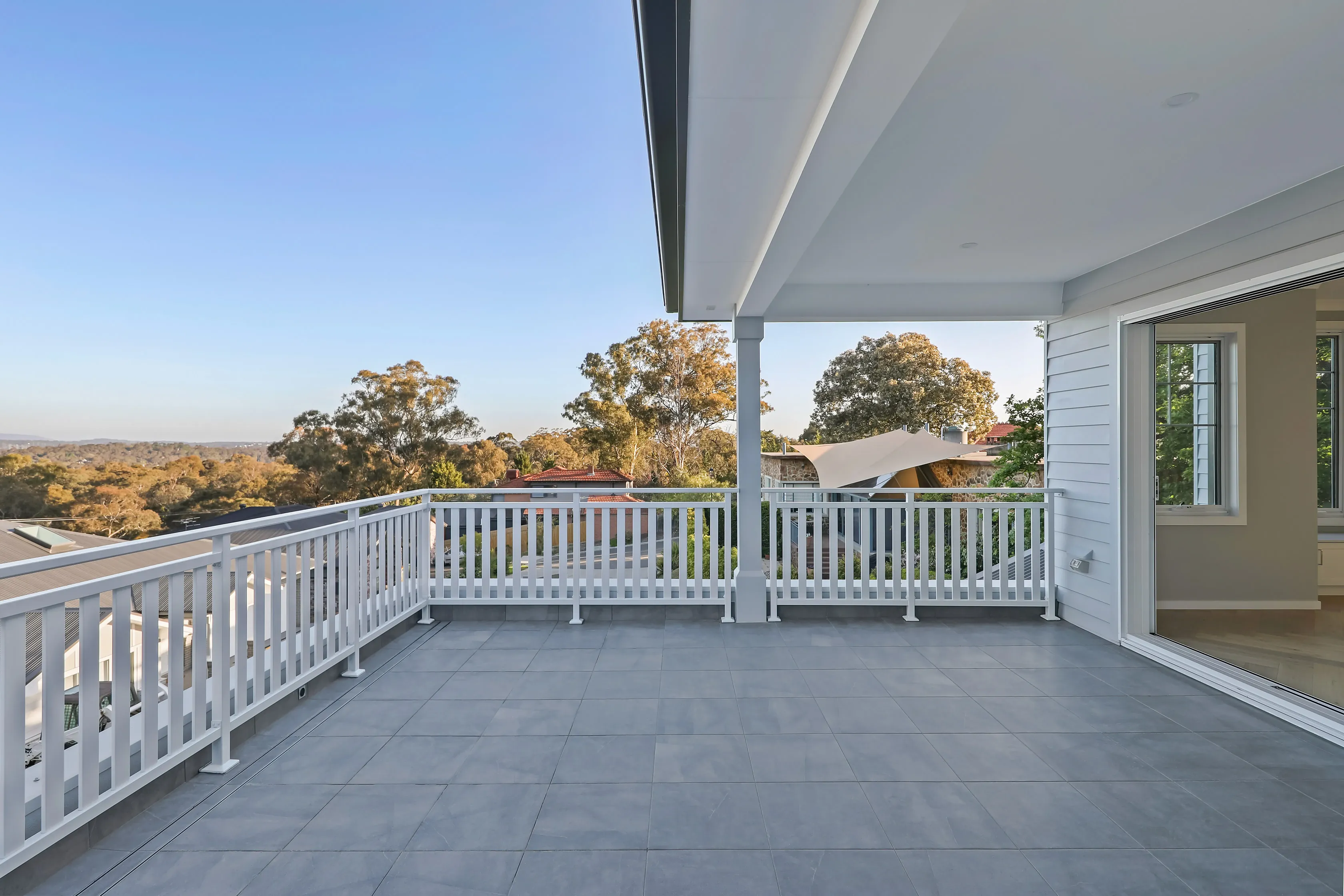 Covered weatherboard balcony with a white timber balustrade, grey tiles and a sliding door, overlooking treetops and rooftops in the valley