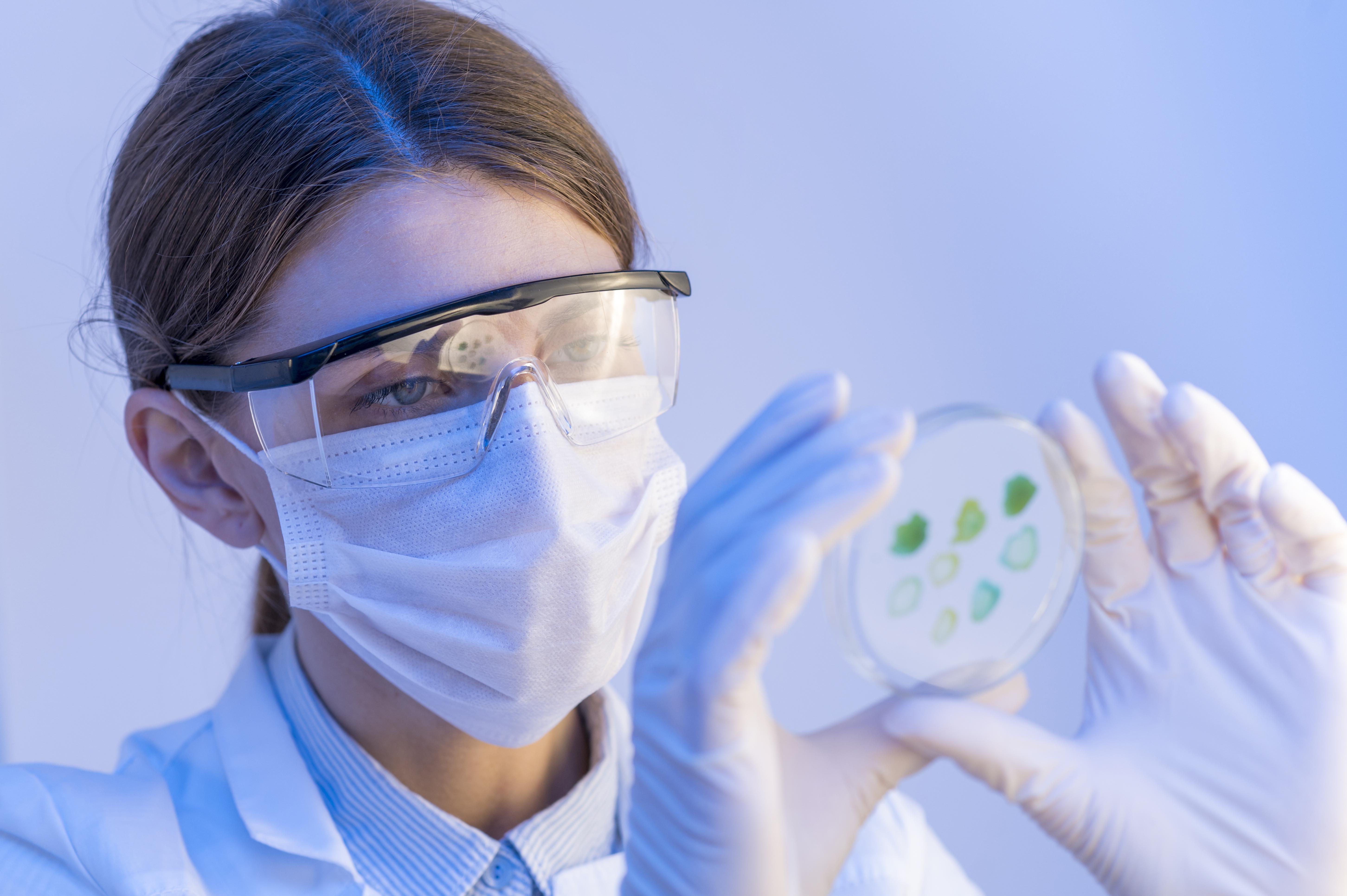 A scientist wearing protective glasses, mask, and gloves examines a petri dish with green samples in a laboratory setting focused on biomanufacturing automation solutions.