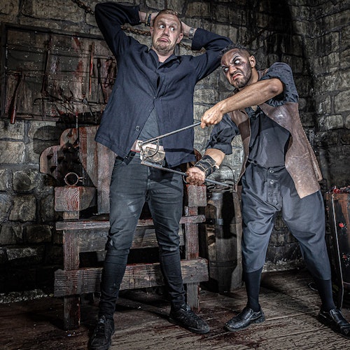 A man restrains another by inserting him into a torture device in a dimly lit, dungeon-like setting.