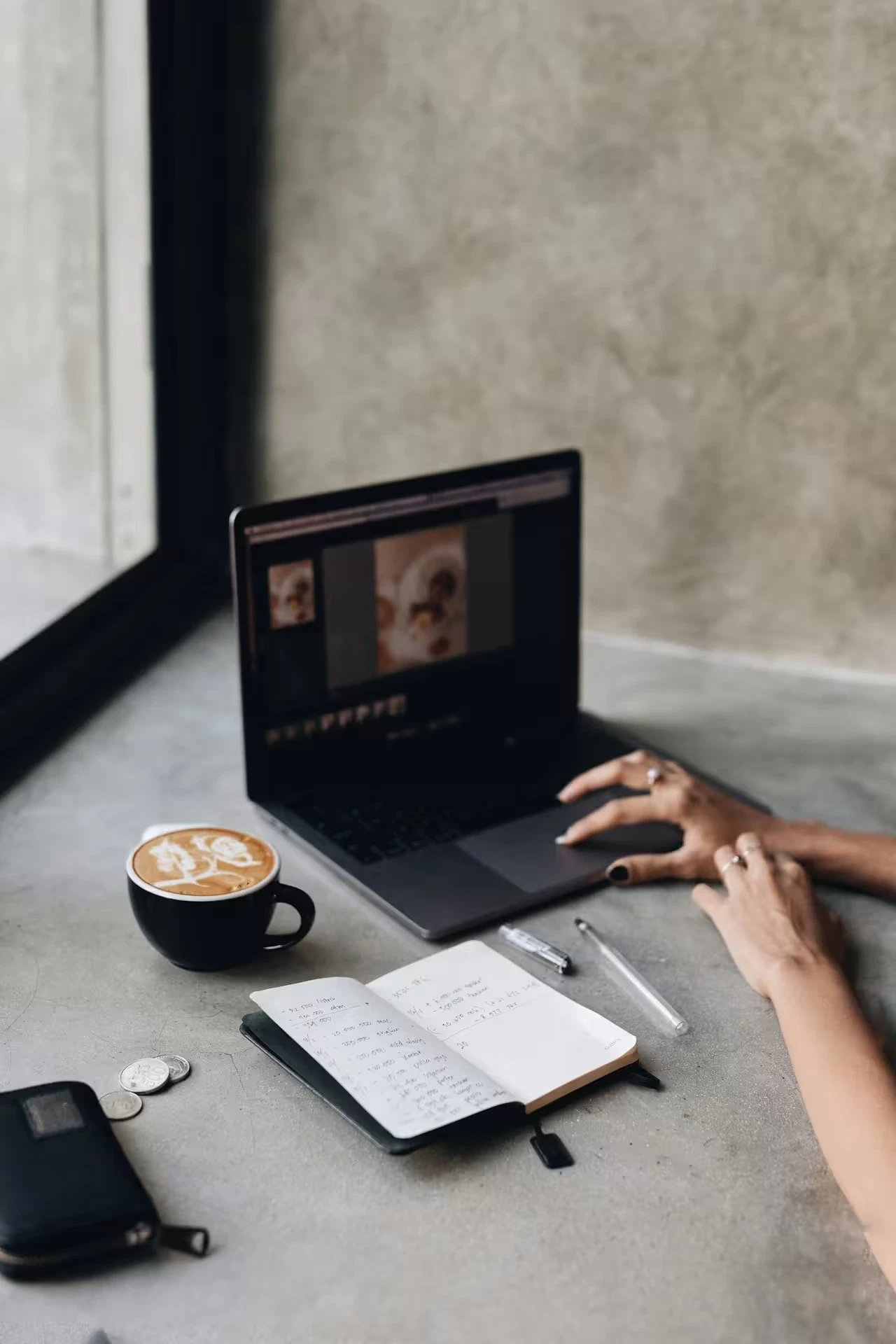 Person typing on a laptop beside an open notebook, a latte, and a small wallet on a concrete surface.