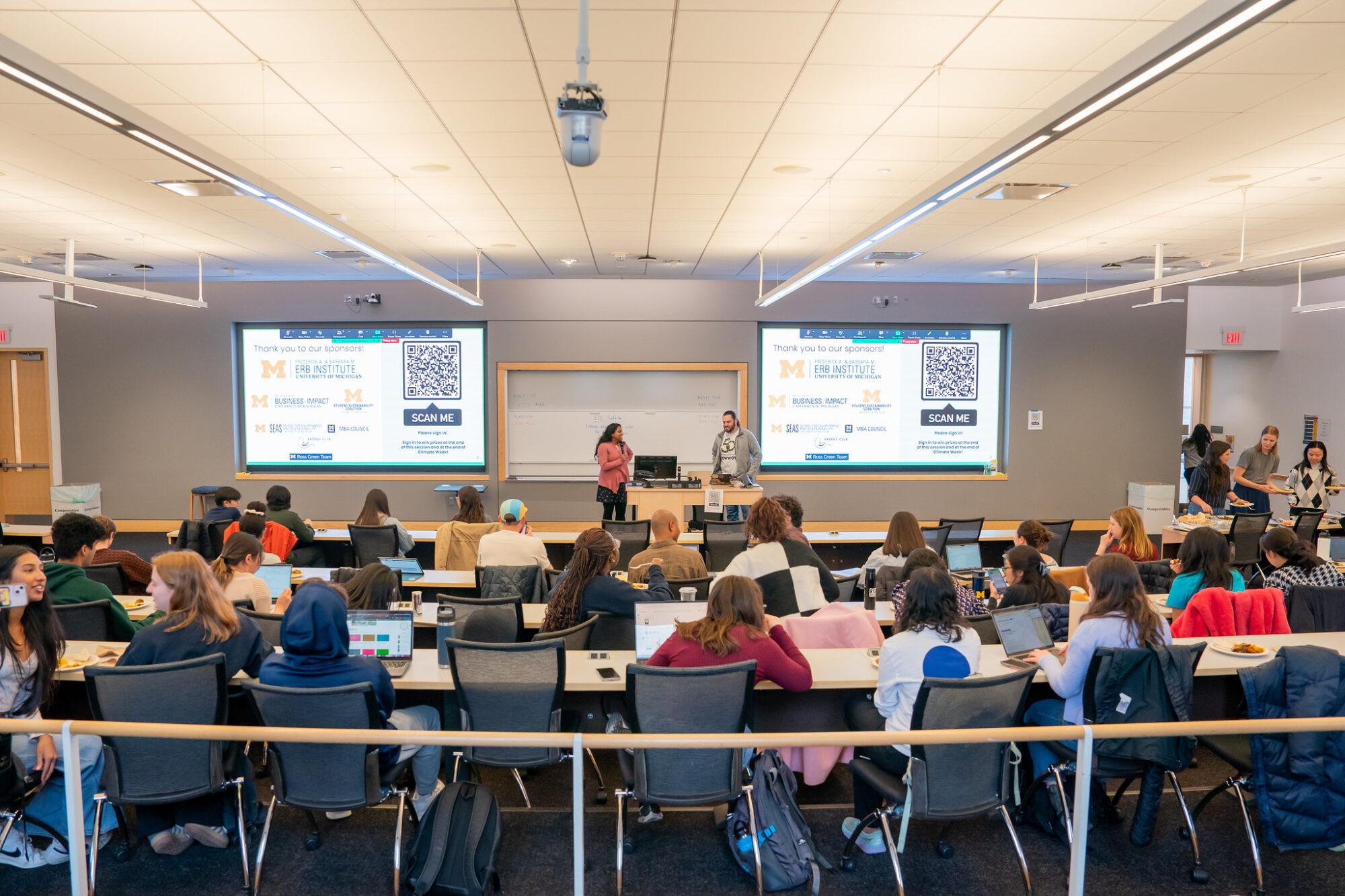 A classroom filled with students attentively listening to a presenter at the front, with screens displaying content.
