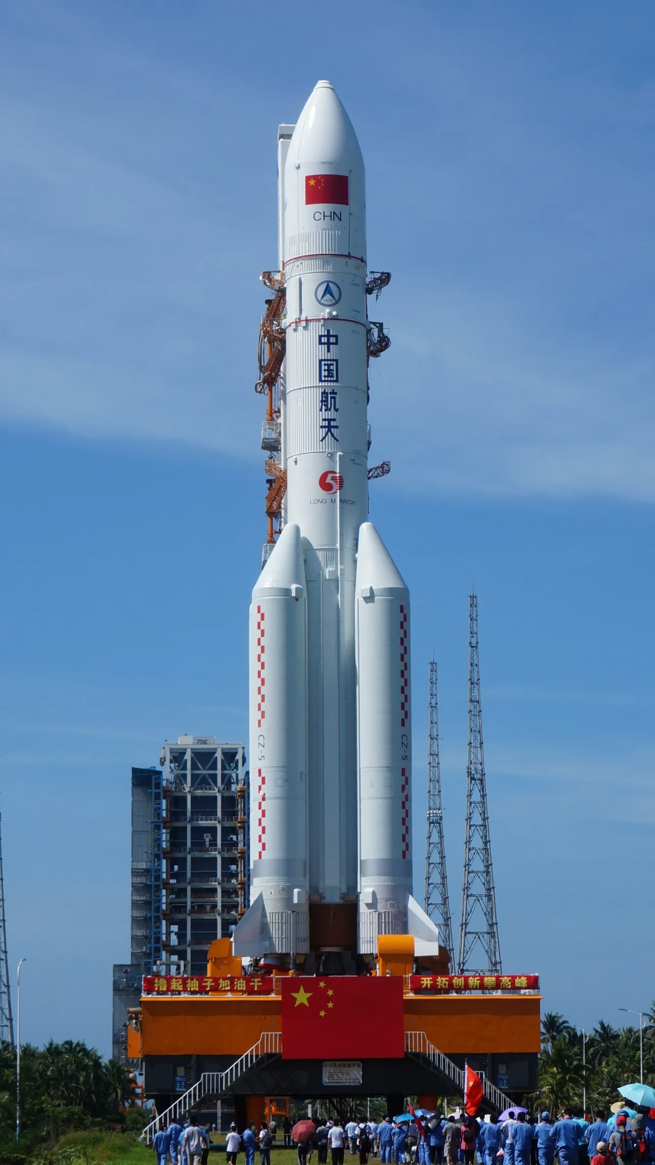 A white Long March 5 rocket stands vertical on a launch pad against a clear blue sky, featuring Chinese flags and surrounded by a crowd of people in blue uniforms, highlighting China's space exploration efforts.