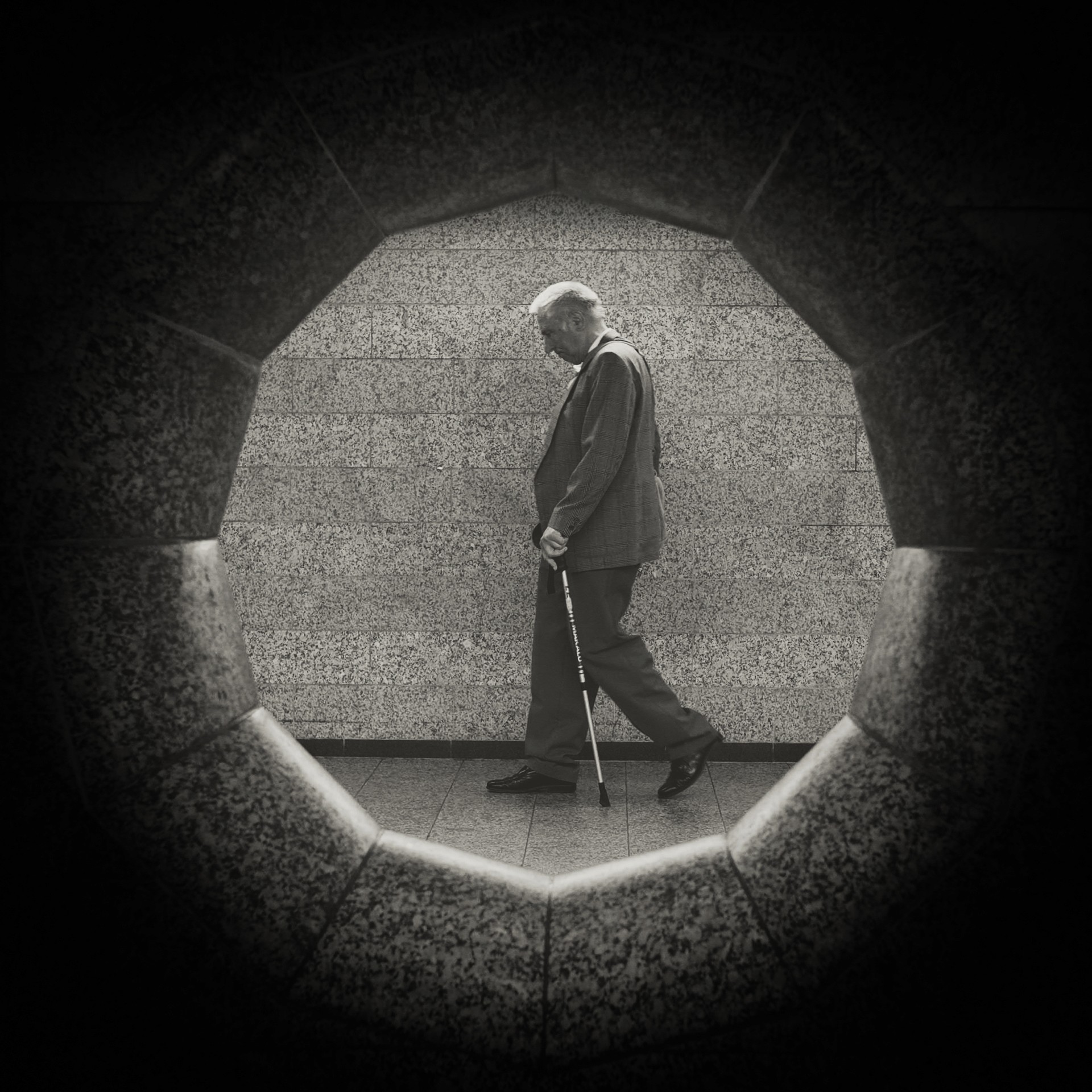Elderly man in suit walking with cane through hallway, viewed through stone archway opening.