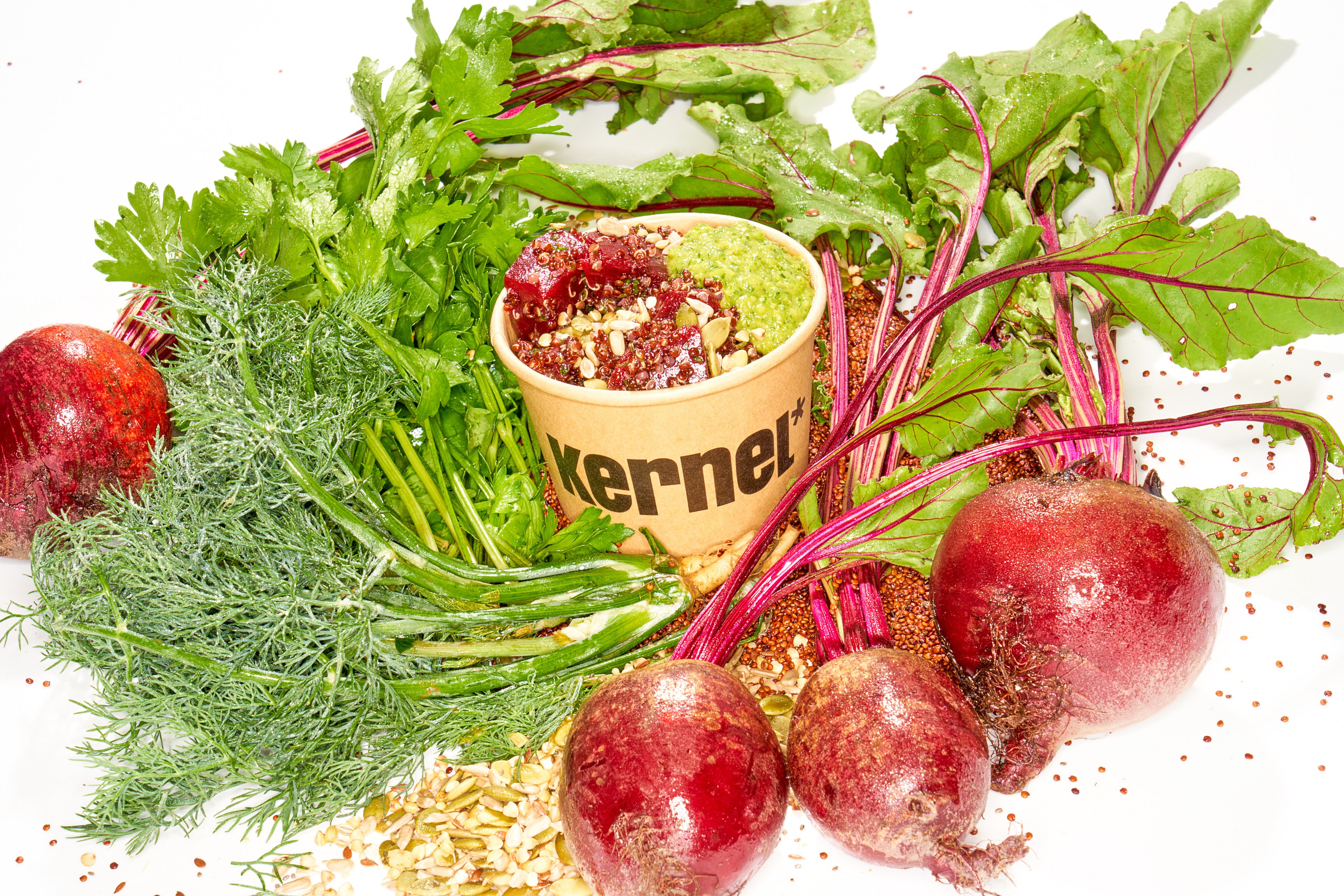 Kernel beet and grain bowl surrounded by fresh beets, herbs, seeds, and greens
