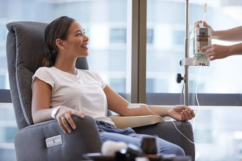 Smiling woman receiving IV infusion therapy in a relaxing clinic setting, a treatment designed to boost hydration, energy, and detoxification from within.