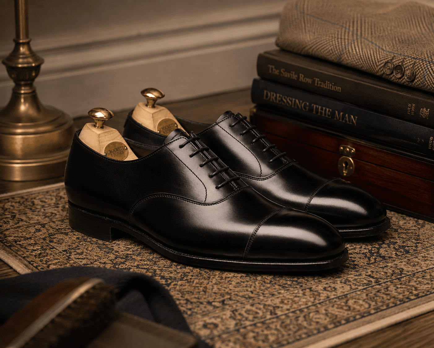 A pair of polished black leather Crockett & Jones cap-toe oxford dress shoes fitted with wooden shoe trees on an ornate patterned rug. The scene includes a brass lamp, stacked menswear books titled "The Savile Row Tradition" and "Dressing the Man", a tweed jacket sleeve, a wooden box, and shoe care brushes in the foreground under warm, classic lighting.