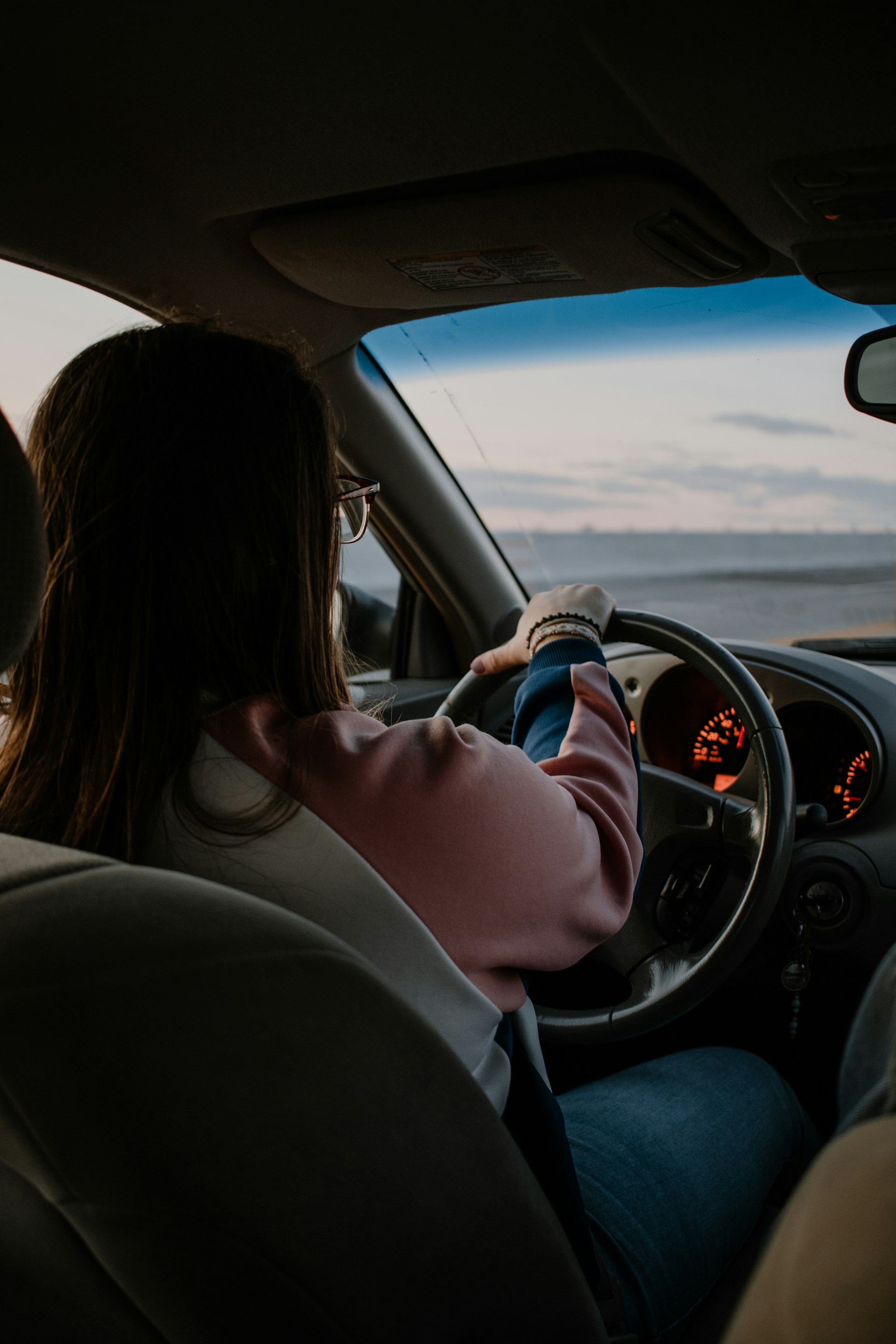  An interior shot from the back seat of a car showing a woman with glasses and long dark hair driving. She is wearing a pink and blue color-blocked jacket and jeans. The dashboard gauges are lit up with an orange glow. Through the windshield, a wide-open landscape, possibly a coastal road with the ocean in the distance, is visible under a dusky sky.