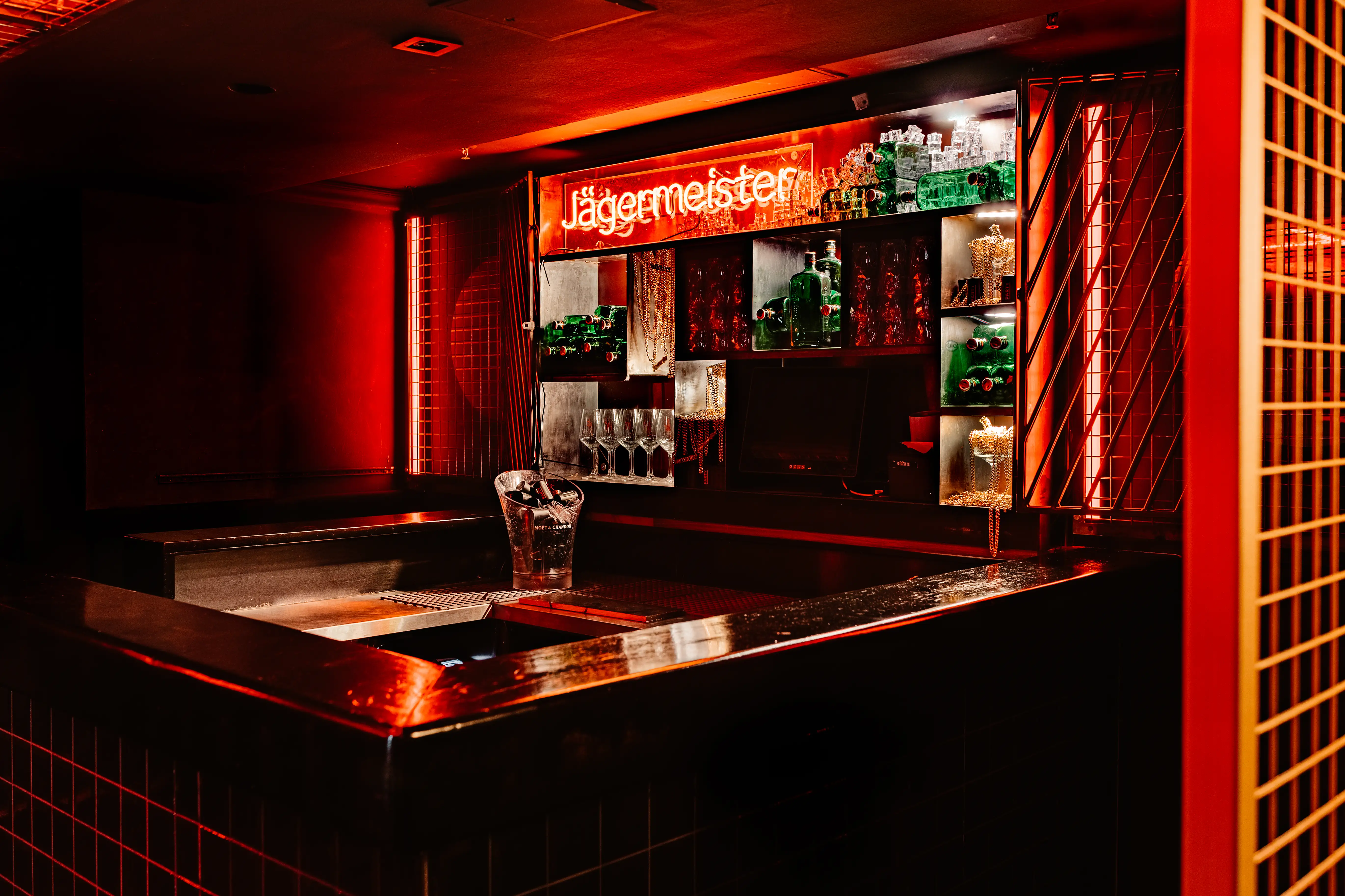 Bar corner at JÄGERMEISTER x CALL ME DRELLA with a copper-toned counter, ice bucket, shelves of green bottles and glasses under an orange Jägermeister neon.