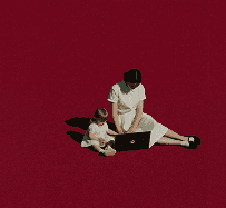 A woman in a white dress sits on the floor with a child, both looking at a small box against a red background.