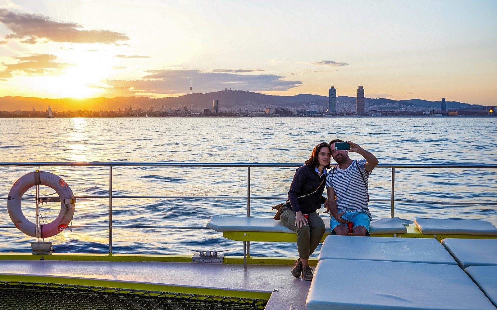Coppia che si fa un selfie durante una crociera in catamarano con lo skyline di Barcellona al tramonto.