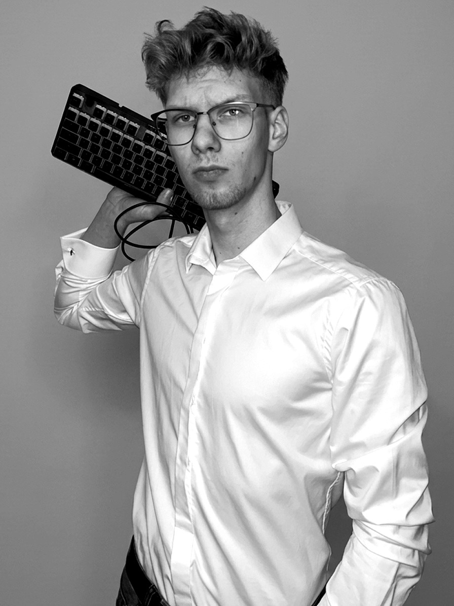 Stanisław 'Stasiowy' Marciniak pozuje do czarno-białego zdjęcia z klawiaturą komputerową opartą o ramię; ma na sobie elegancką białą koszulę i okulary.