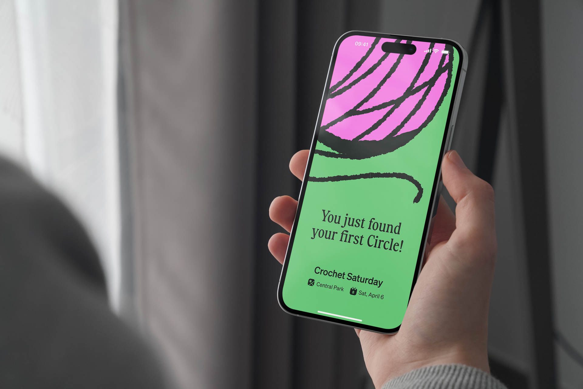 A hand holds a smartphone displaying a bright green screen with a stylized pink flower graphic and the text "You just found your first Circle!" in black. Below the graphic, the text "Crochet Saturday" is visible, along with "Central Park" and "Sat., Apr. 6". The phone’s top shows the time as 08:41. The background is blurred, with a light-colored curtain visible to the left.