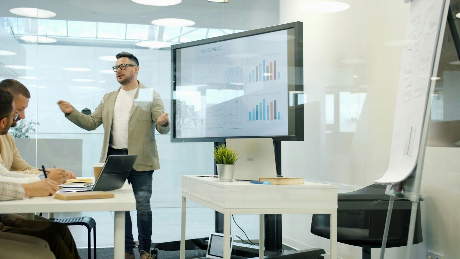 A man presenting sales data to a small team in a modern glass walled office using a large digital screen and collaborative workspace setup.