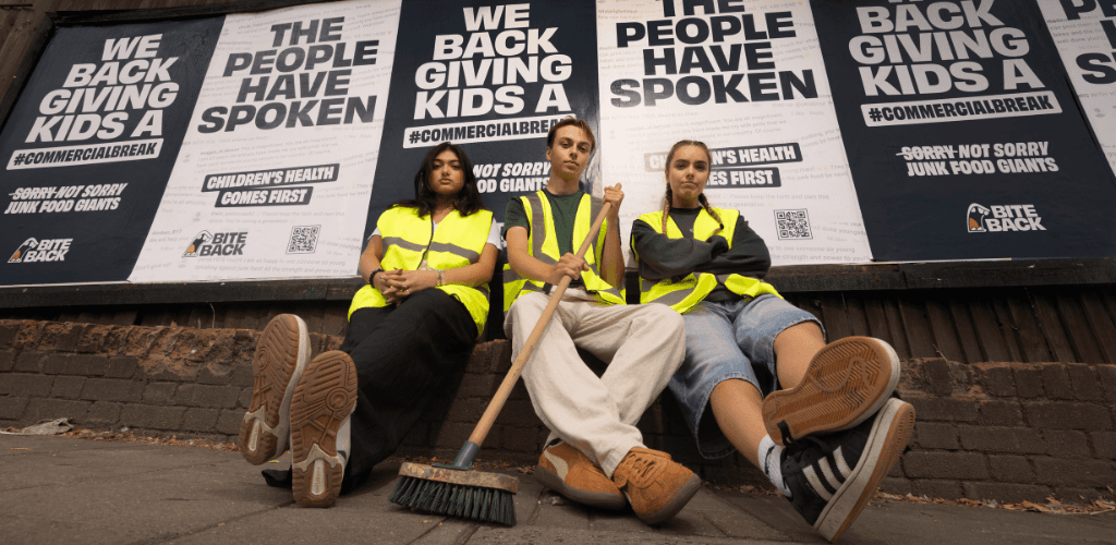 photo of young people wearing yellow jackets, one of them holding a broom. Behind them poster showing sentences: 'we back giving kids a #commercialbreak, ' the people have spoken'
