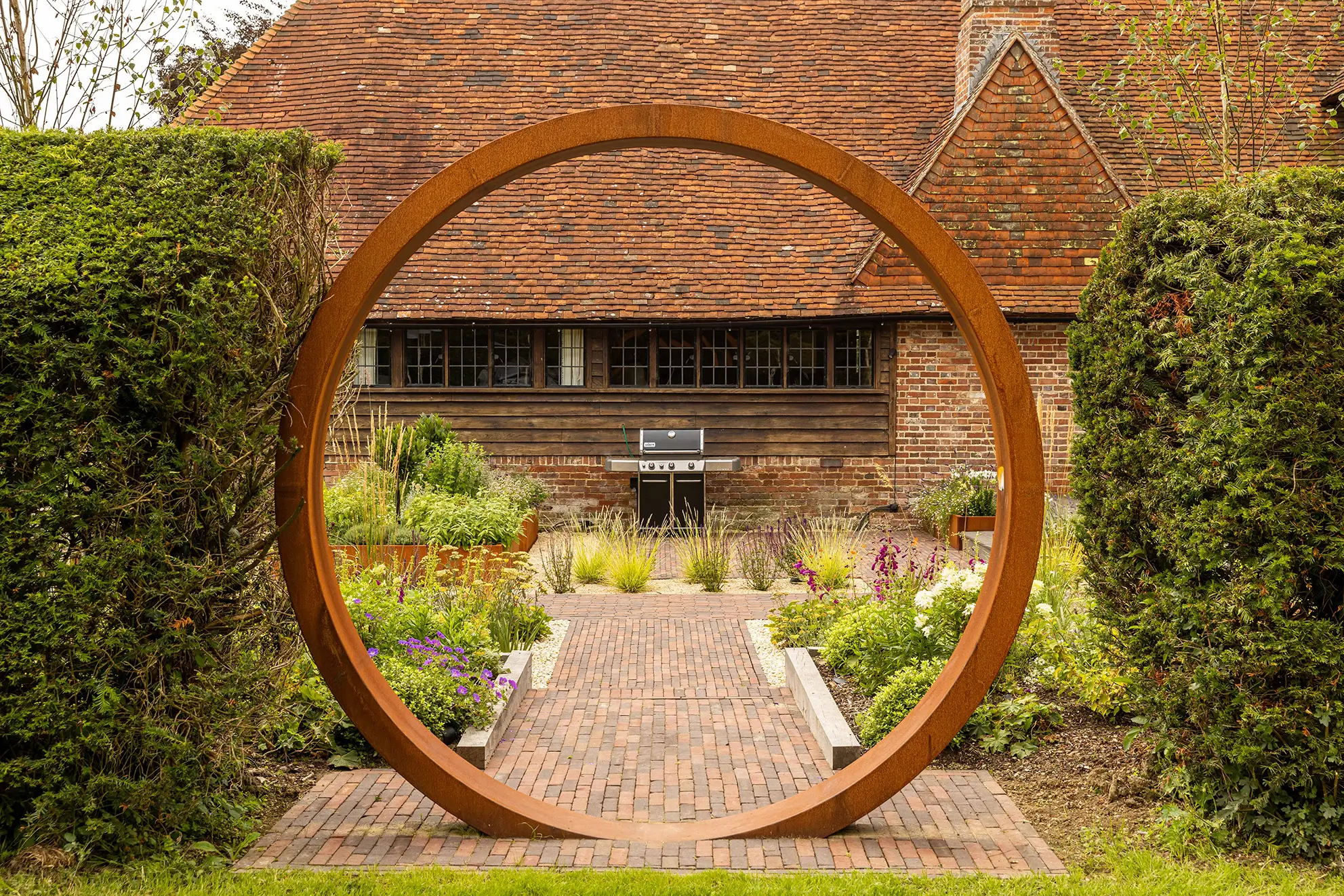 A circular wooden arch framed by greenery leads to a pathway and a rustic building in the background.