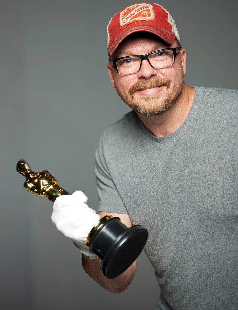 A man in glasses and a red cap smiles at the camera while holding an Oscar statuette in a white glove against a gray studio backdrop.